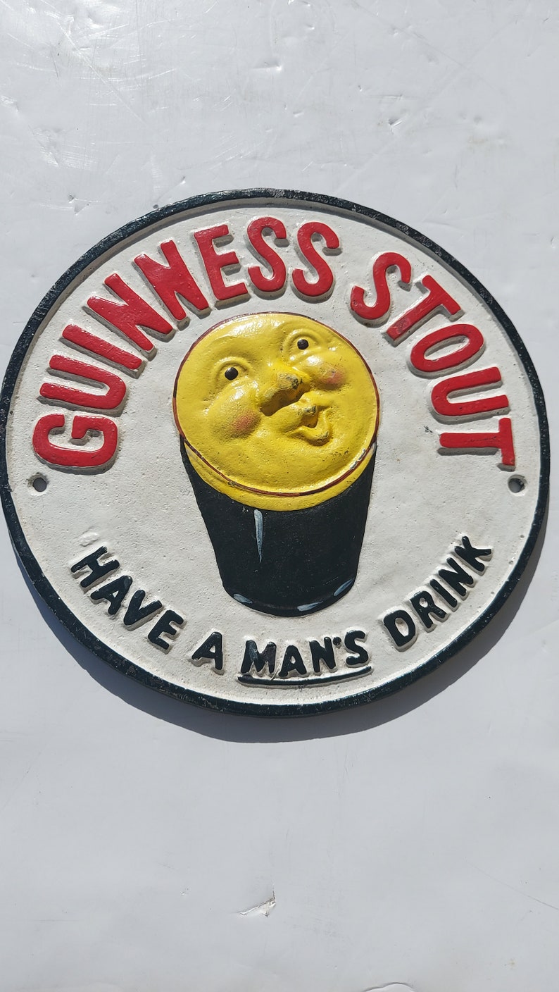 May include: A white, round, cast iron sign with a black border. The sign has red lettering that says "Guinness Stout" and "Have a Man's Drink". In the center of the sign is a yellow cartoon face with a black pint glass.