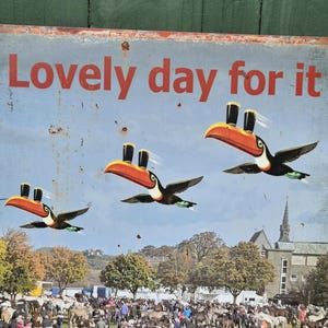 May include: A vintage metal sign with the text "Lovely day for it" in red. The sign features three toucans with orange beaks and top hats flying over a crowd of people and horses in a field.
