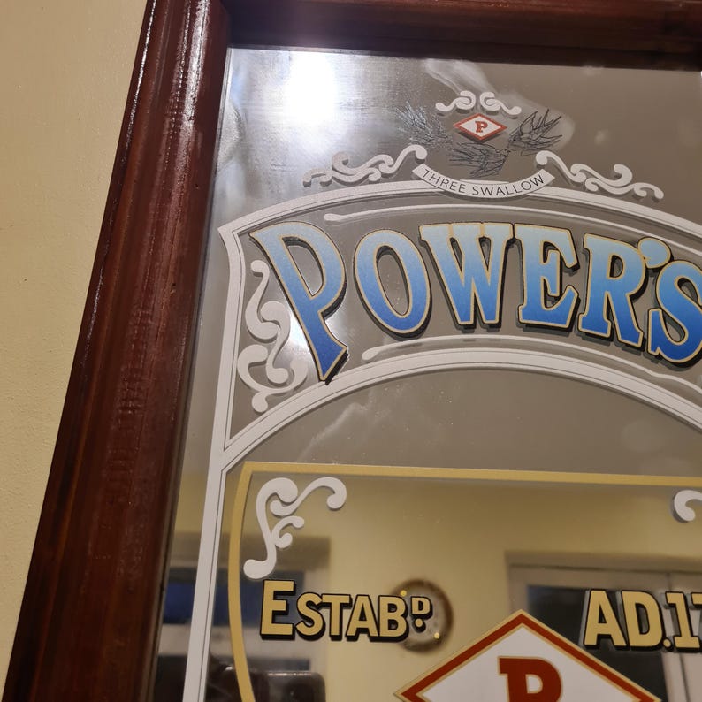 Puede incluir: Un espejo vintage con marco de madera marr&oacute;n oscuro. El espejo presenta la palabra "POWER'S" en una fuente azul y blanca, con las palabras "THREE SWALLOW" encima. El espejo tambi&eacute;n tiene la palabra "ESTAB" y "AD17" en dorado.