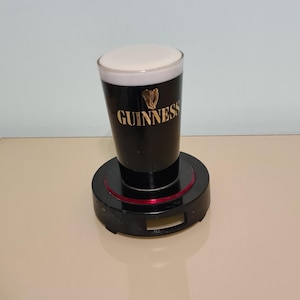 May include: A black and gold Guinness pint glass with a white head of foam sits on a black circular base with a red ring around the edge.