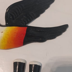 Op de afbeelding: Een geschilderde vogeldecoratie met zwarte vleugels, een gele en rode body en witte accenten. De vogel is een decoratief item, mogelijk voor een muur of tuin.