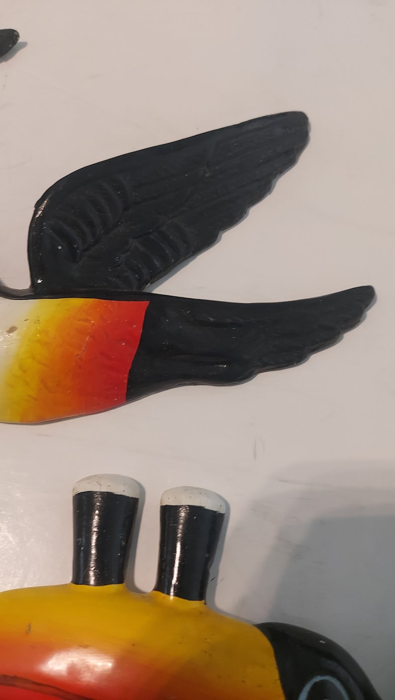 Op de afbeelding: Een geschilderde vogeldecoratie met zwarte vleugels, een gele en rode body en witte accenten. De vogel is een decoratief item, mogelijk voor een muur of tuin.