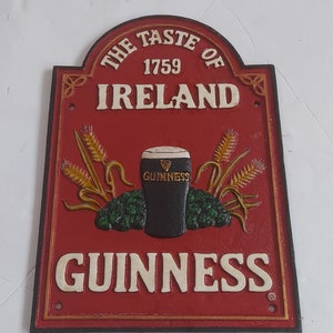 May include: Red and gold metal sign with the text "THE TASTE OF IRELAND 1759 GUINNESS". The sign features a pint of Guinness, wheat stalks, and hops.