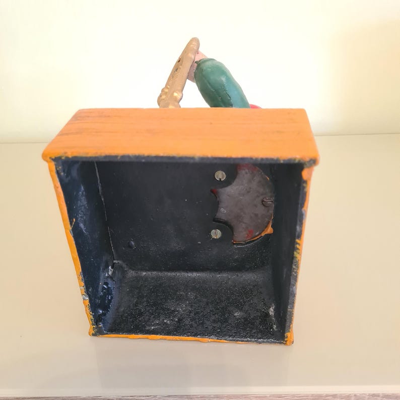 May include: Vintage orange and black cast iron bank with a painted figure holding a baseball bat. The bank has a slot on top and a coin release mechanism on the side. The figure is wearing a green shirt and red hat.