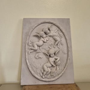 Op de afbeelding: Een rechthoekige lichtgrijze wandplaat met een ovaal reliëf van drie gevleugelde cherubijnen te midden van wolken. De centrale figuren zijn afgebeeld in een klassieke stijl met gedetailleerde vleugels en zachte gelaatstrekken.