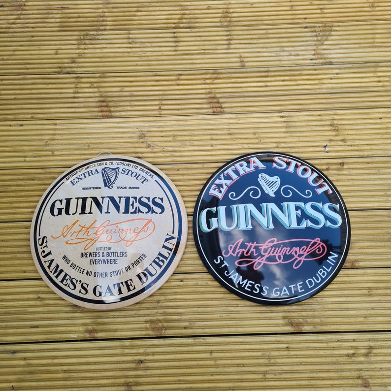May include: Two round Guinness signs. One is cream-colored with black and orange text, the other is black with blue and pink text. Both signs feature the Guinness logo and the words "Extra Stout."