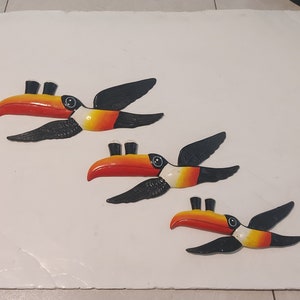 Op de afbeelding: Drie wanddecoraties van toekans. Elke toekan heeft een rode en oranje snavel, zwarte vleugels en een wit en geel lichaam. De toekans zijn diagonaal op een wit oppervlak geplaatst.