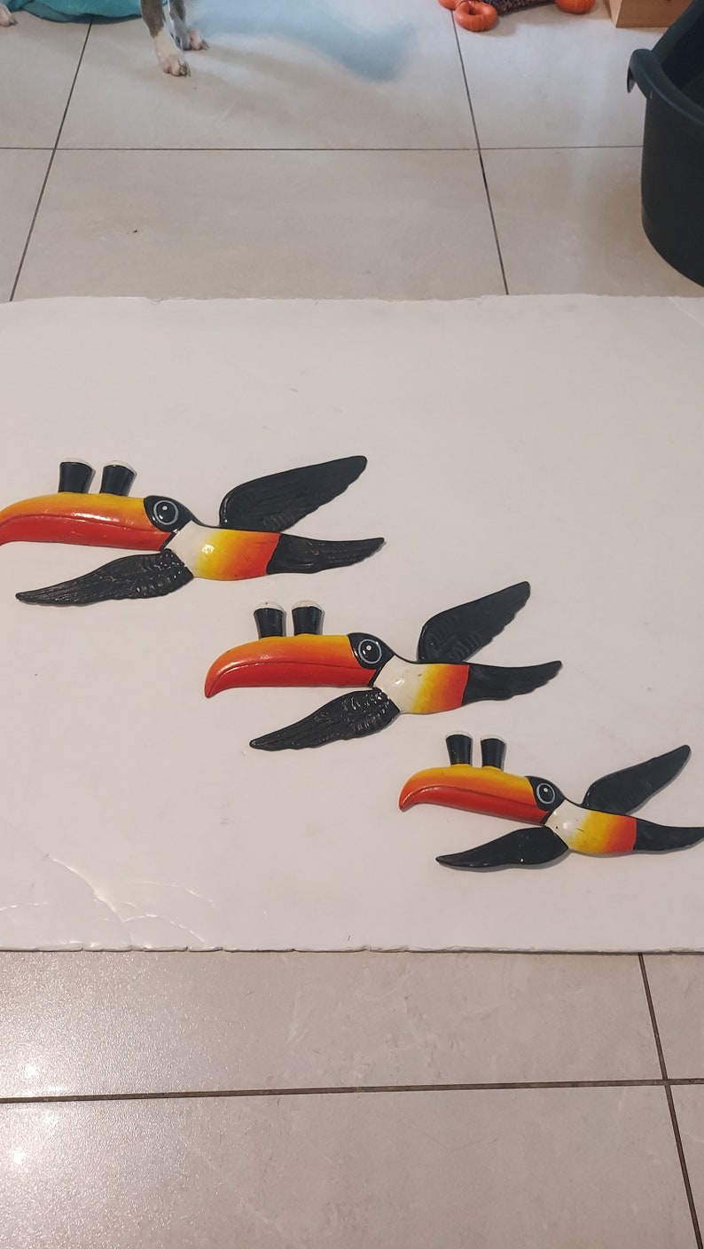 Op de afbeelding: Drie wanddecoraties van toekans. Elke toekan heeft een rode en oranje snavel, zwarte vleugels en een wit en geel lichaam. De toekans zijn diagonaal op een wit oppervlak geplaatst.