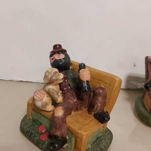 May include: A ceramic figurine of a man with a beard and a hat sitting on a bench with a dog. The man is wearing a red and green shirt and brown pants. The dog is brown and white. The bench is yellow and brown. The figurine is sitting on a green base.