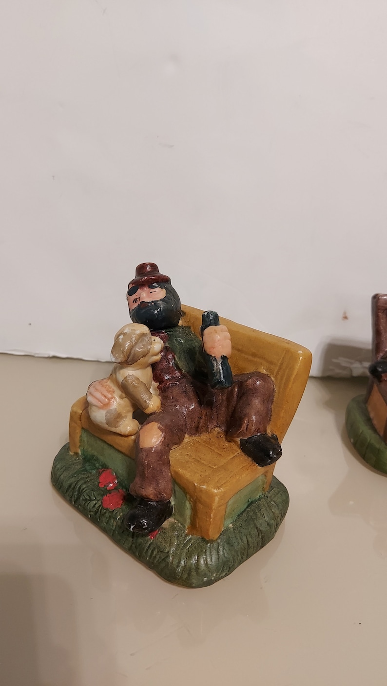 May include: A ceramic figurine of a man with a beard and a hat sitting on a bench with a dog. The man is wearing a red and green shirt and brown pants. The dog is brown and white. The bench is yellow and brown. The figurine is sitting on a green base.
