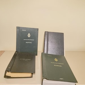 Könnte beinhalten: Vier dunkelgrüne und schwarze Ordner mit der Aufschrift The Royal Ulster Constabulary Manual. Diese rechteckigen Handbücher haben ein goldenes Emblem auf den Vorderseiten, Metallbindemechanismen und sichtbare Papierseiten in den Einbänden.