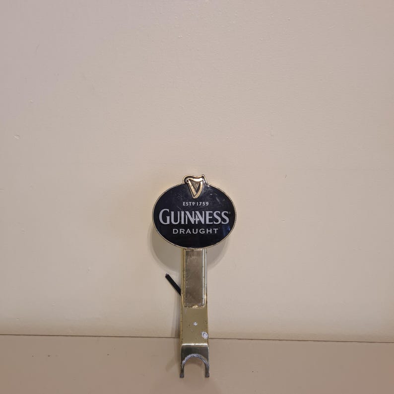 May include: A Guinness Draught tap handle with a black and gold oval emblem. The emblem features the Guinness logo and the words "Guinness Draught". The handle is gold-colored and has a curved bottom.