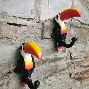 Könnte beinhalten: Zwei schwarze Wandhaken in Toukanform mit gelben und roten Schnäbeln. Die Haken sind an einer Steinwand befestigt.