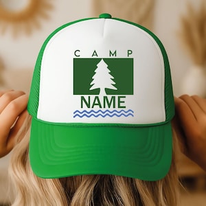 May include: A green and white trucker hat with the word "CAMP" above a green rectangle with a tree graphic. Below the tree is the word "NAME" above blue wavy lines. The hat is being held by a person.