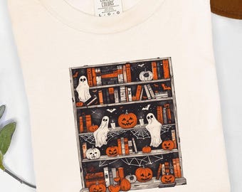 May include: Cream-colored t-shirt featuring a Halloween-themed graphic. The graphic depicts a bookshelf filled with books, pumpkins, ghosts, and bats. The shirt has a crew neck and short sleeves. The brand label "Comfort Colors" is visible.