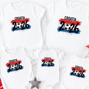 以下が含まれることがあります： サッカーボールのグラフィックの上に「CROATIA 2026」の文字が入った白いTシャツ。デザインは赤、白、青の色を使用し、使い古されたペイント効果があります。Tシャツは様々なサイズがあります。