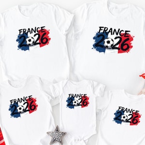 K&ouml;nnte beinhalten: Wei&szlig;e T-Shirts mit einem Fu&szlig;ball-Grafik und dem Text "FRANCE 2026" in Schwarz. Die Grafik enth&auml;lt die Farben der franz&ouml;sischen Flagge: Blau, Wei&szlig; und Rot. Die Shirts sind in verschiedenen Gr&ouml;&szlig;en erh&auml;ltlich, darunter ein Body.