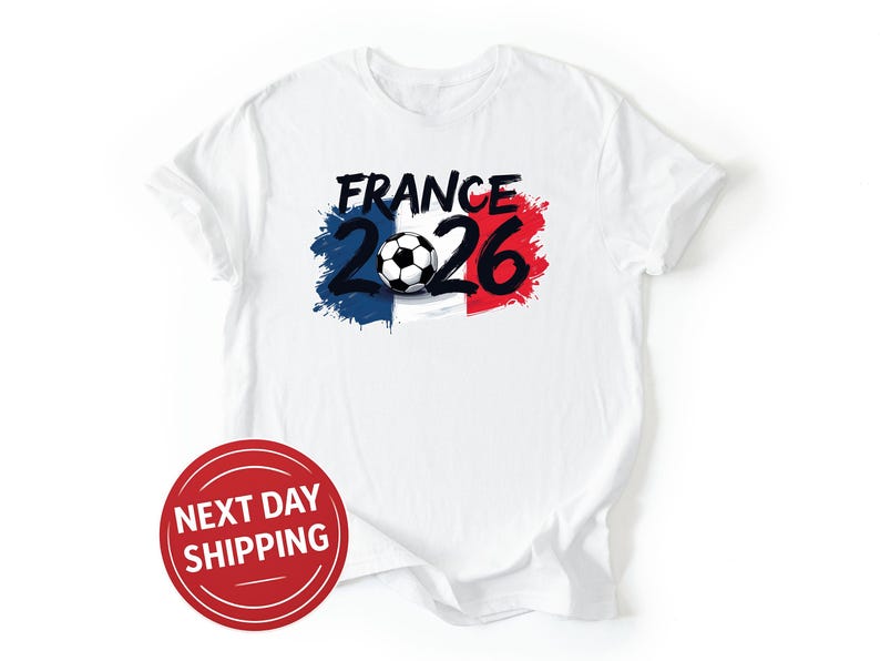 May include: White t-shirt featuring the text "FRANCE 2026" in black, with a soccer ball graphic. The design incorporates the French flag colors: blue, white, and red. A red circle with the text "NEXT DAY SHIPPING" is in the lower left.