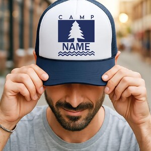 May include: A navy blue and white trucker hat. The front panel displays the word "CAMP" above a navy blue rectangle with a white pine tree and the word "NAME" below. The hat has a curved peak and a mesh back.