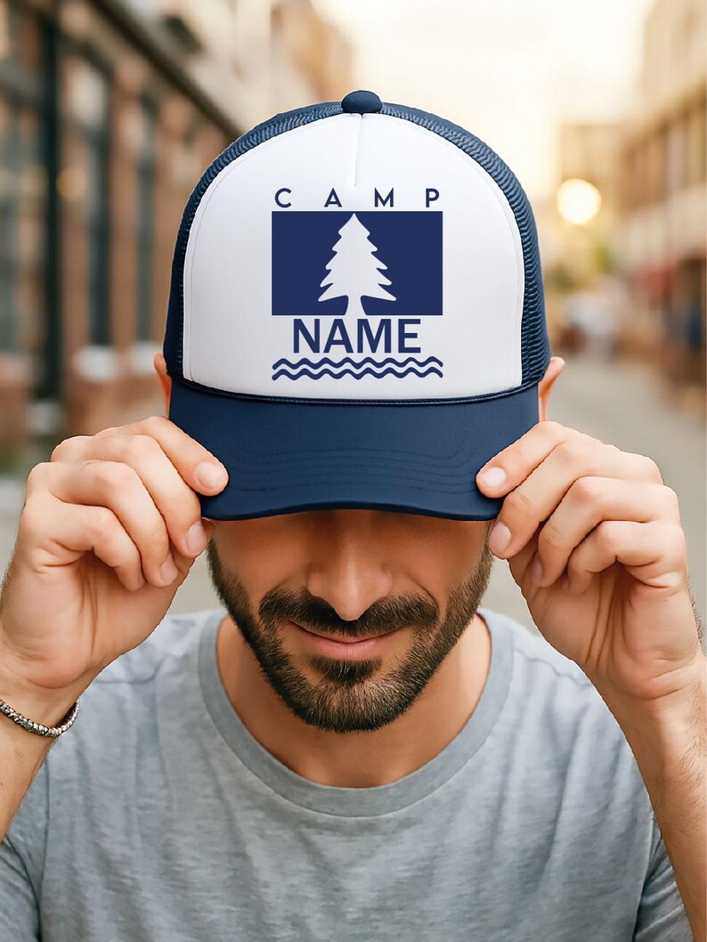 May include: A navy blue and white trucker hat. The front panel displays the word "CAMP" above a navy blue rectangle with a white pine tree and the word "NAME" below. The hat has a curved peak and a mesh back.