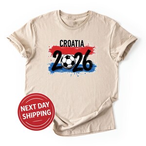 以下が含まれることがあります： ベージュのTシャツ。黒字で「CROATIA」の文字が赤と青のペイントストロークデザインの上に書かれています。デザインの下には、サッカーボールを中央に配置した黒字の数字「2026」があります。左下には「NEXT DAY SHIPPING」と書かれた赤い円があります。