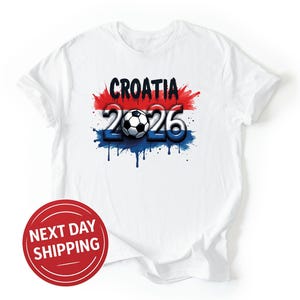 May include: White t-shirt with a graphic design featuring the word "CROATIA" in black, the number "2026", and a soccer ball. The design incorporates red, white, and blue colors, with a dripping paint effect. A red circle with "NEXT DAY SHIPPING" is in the lower left.