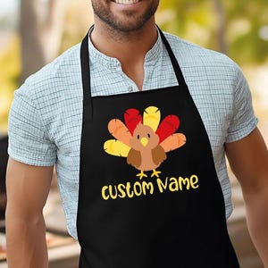 May include: Black apron featuring a cartoon turkey with red, orange, and yellow feathers. The text "custom Name" is in yellow script below the turkey. The apron is worn over a light blue plaid shirt.