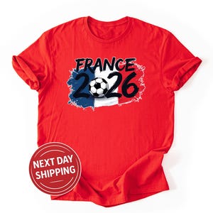K&ouml;nnte beinhalten: Rotes T-Shirt mit einem Grafikdesign. Das Design enth&auml;lt das Wort "FRANCE" in Schwarz, die Zahl "26" und einen Fu&szlig;ball. Das Design integriert die Farben der franz&ouml;sischen Flagge: Blau, Wei&szlig; und Rot. Rundhalsausschnitt.