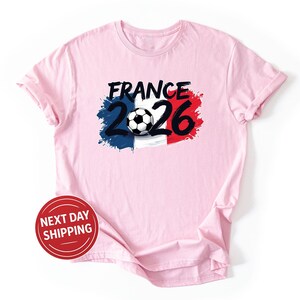 K&ouml;nnte beinhalten: Hellrosa T-Shirt mit einem Grafikdesign. Das Design beinhaltet das Wort "FRANCE" in Schwarz, die Zahlen "2026" und einen Fu&szlig;ball. Das Design integriert die Farben der franz&ouml;sischen Flagge: Blau, Wei&szlig; und Rot.