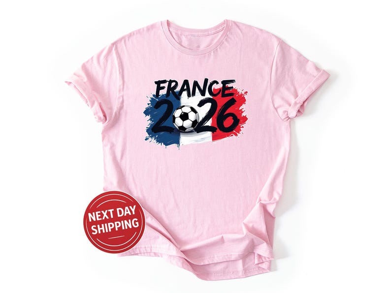 K&ouml;nnte beinhalten: Hellrosa T-Shirt mit einem Grafikdesign. Das Design beinhaltet das Wort "FRANCE" in Schwarz, die Zahlen "2026" und einen Fu&szlig;ball. Das Design integriert die Farben der franz&ouml;sischen Flagge: Blau, Wei&szlig; und Rot.