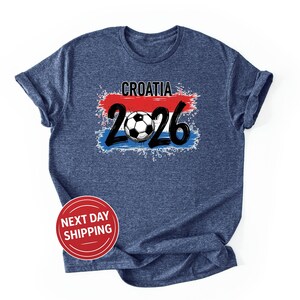 以下が含まれることがあります： グラフィックデザインのネイビーブルーTシャツ。デザインには、数字「2026」の上に「CROATIA」という単語とサッカーボールが含まれています。デザインにはクロアチア国旗の色が組み込まれています。「NEXT DAY SHIPPING」という言葉が入った赤い円が下にあります。
