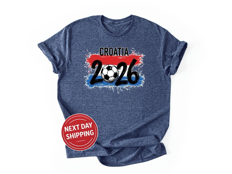 以下が含まれることがあります： グラフィックデザインのネイビーブルーTシャツ。デザインには、数字「2026」の上に「CROATIA」という単語とサッカーボールが含まれています。デザインにはクロアチア国旗の色が組み込まれています。「NEXT DAY SHIPPING」という言葉が入った赤い円が下にあります。