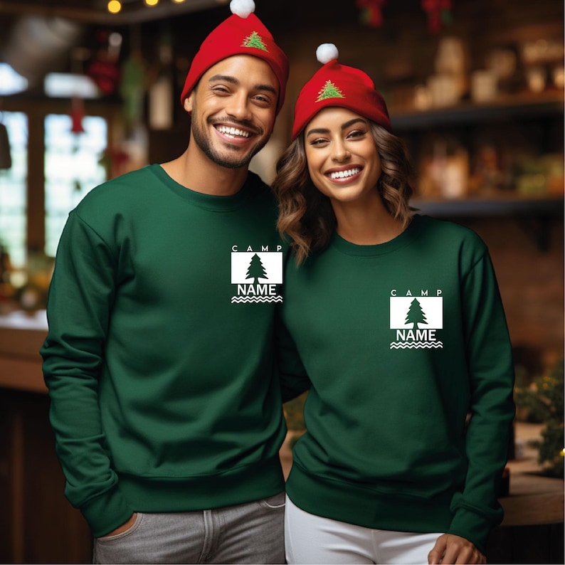 Puede incluir: Dos personas con sudaderas verdes a juego y gorros rojos. Las sudaderas tienen un gr&aacute;fico blanco de un &aacute;rbol con la palabra "CAMP" encima y "NAME" debajo. Los gorros tienen un &aacute;rbol verde bordado.
