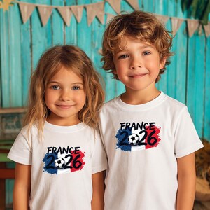 K&ouml;nnte beinhalten: Zwei Kinder tragen wei&szlig;e T-Shirts mit dem Text "FRANCE 2026" in Schwarz, einem Fu&szlig;ball und den Farben der franz&ouml;sischen Flagge. Die T-Shirts sind kurz&auml;rmlig.