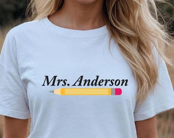 May include: White t-shirt with the name "Mrs. Anderson" in black script above a yellow pencil graphic with a gray tip and a pink eraser. The shirt is made of a soft material.