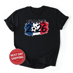 K&ouml;nnte beinhalten: Schwarzes T-Shirt mit einem Fu&szlig;ball und dem Text "France 2026". Das Design verwendet die Farben der franz&ouml;sischen Flagge: Blau, Wei&szlig; und Rot. Ein roter Kreis mit dem Text "NEXT DAY SHIPPING" ist ebenfalls sichtbar.