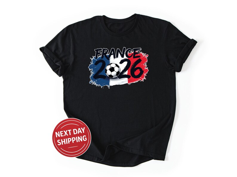 K&ouml;nnte beinhalten: Schwarzes T-Shirt mit einem Fu&szlig;ball und dem Text "France 2026". Das Design verwendet die Farben der franz&ouml;sischen Flagge: Blau, Wei&szlig; und Rot. Ein roter Kreis mit dem Text "NEXT DAY SHIPPING" ist ebenfalls sichtbar.