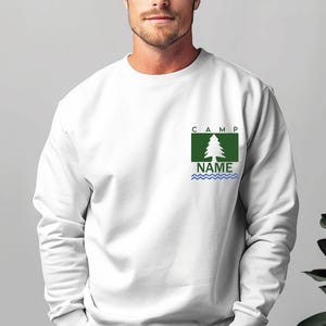Puede incluir: Sudadera blanca con un dise&ntilde;o gr&aacute;fico verde y azul. El dise&ntilde;o incluye un &aacute;rbol, la palabra "CAMP" encima del &aacute;rbol y la palabra "NAME" debajo, con l&iacute;neas onduladas azules. La sudadera es de manga larga.