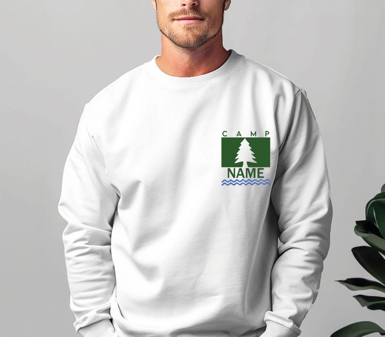 Puede incluir: Sudadera blanca con un dise&ntilde;o gr&aacute;fico verde y azul. El dise&ntilde;o incluye un &aacute;rbol, la palabra "CAMP" encima del &aacute;rbol y la palabra "NAME" debajo, con l&iacute;neas onduladas azules. La sudadera es de manga larga.