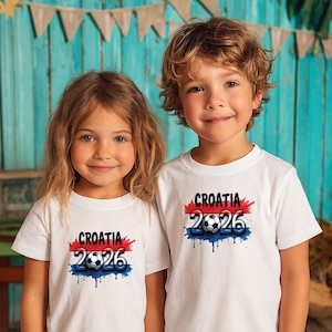 May include: Two children wearing white t-shirts with the text "CROATIA 2026" in red, white, and blue, with a soccer ball graphic. The design has a distressed, paint-splatter effect. The children are smiling.