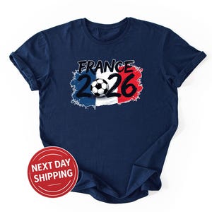 K&ouml;nnte beinhalten: Marineblaues T-Shirt mit einem Aufdruck "FRANCE 2026" &uuml;ber einem Fu&szlig;ball, vor dem Hintergrund der franz&ouml;sischen Flagge. Das Design ist in Wei&szlig;, Rot und Blau gehalten. Ein roter Kreis mit dem Text "NEXT DAY SHIPPING" befindet sich unten links.