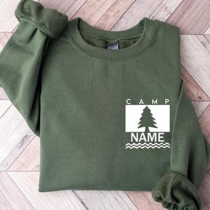 Puede incluir: Una sudadera verde oliva con la palabra "CAMP" encima de un rect&aacute;ngulo blanco que contiene un dibujo de un &aacute;rbol y la palabra "NAME" debajo. La sudadera tiene mangas largas y cuello redondo.
