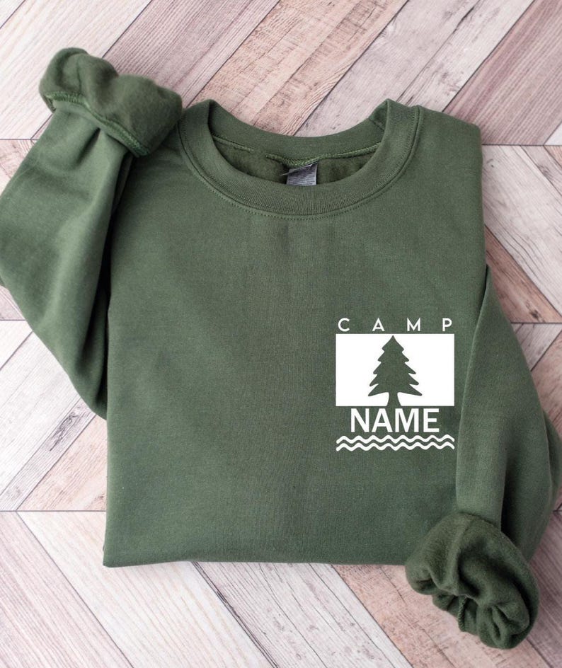 Puede incluir: Una sudadera verde oliva con la palabra "CAMP" encima de un rect&aacute;ngulo blanco que contiene un dibujo de un &aacute;rbol y la palabra "NAME" debajo. La sudadera tiene mangas largas y cuello redondo.