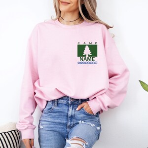 Puede incluir: Una sudadera rosa claro con un dise&ntilde;o de &aacute;rbol verde y blanco, la palabra "CAMP" encima del &aacute;rbol y "NAME" debajo, con l&iacute;neas onduladas azules. La sudadera se usa con vaqueros azules.