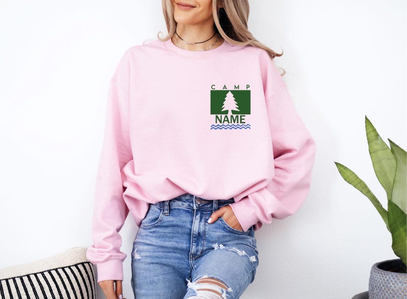 Puede incluir: Una sudadera rosa claro con un dise&ntilde;o de &aacute;rbol verde y blanco, la palabra "CAMP" encima del &aacute;rbol y "NAME" debajo, con l&iacute;neas onduladas azules. La sudadera se usa con vaqueros azules.