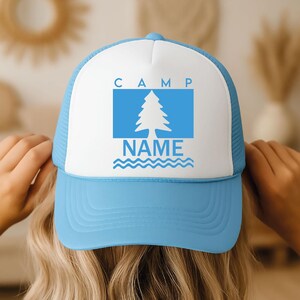 May include: A light blue and white trucker hat. The front panel has the word "CAMP" above a blue rectangle with a white tree graphic. The word "NAME" is below the tree, with blue wavy lines. The hat has a mesh back.