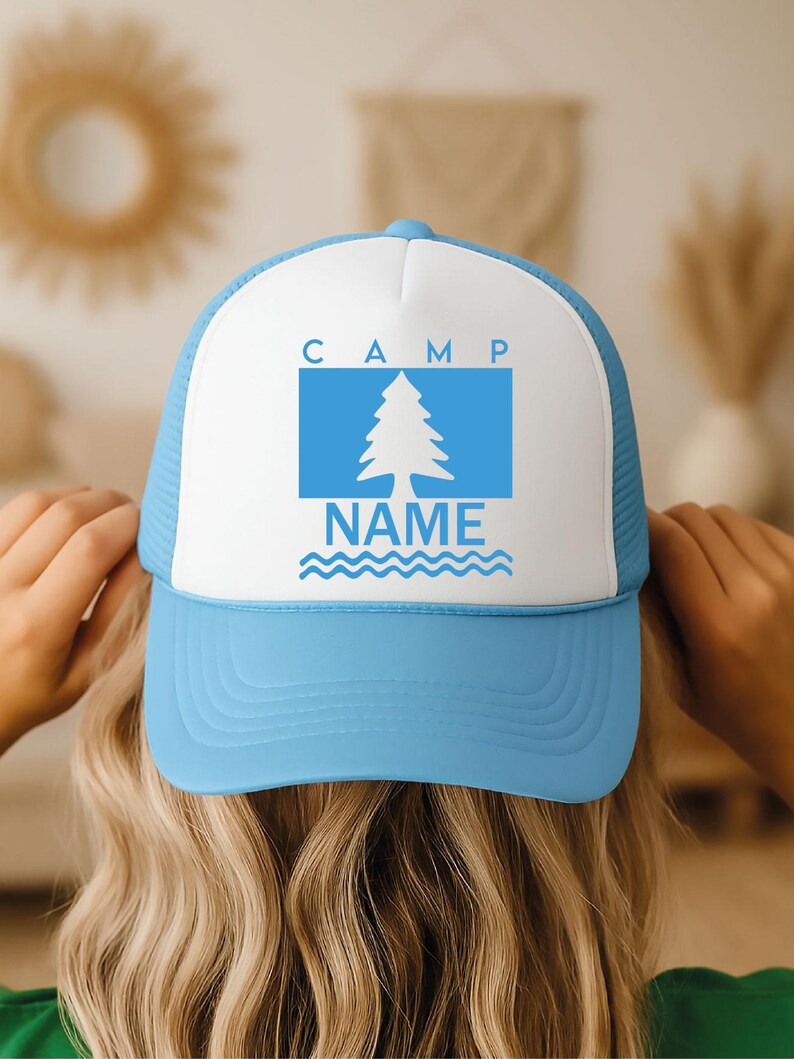 May include: A light blue and white trucker hat. The front panel has the word "CAMP" above a blue rectangle with a white tree graphic. The word "NAME" is below the tree, with blue wavy lines. The hat has a mesh back.