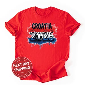 May include: A red t-shirt featuring the word "CROATIA" above the numbers "2026" with a soccer ball in the center. The design incorporates red, white, and blue colors, with a dripping paint effect. The shirt is made of a soft material.