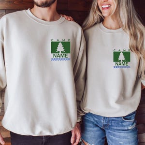 Puede incluir: Dos sudaderas beige. Cada una tiene un dise&ntilde;o gr&aacute;fico cuadrado verde con un &aacute;rbol blanco, la palabra "CAMP" arriba y "NAME" con l&iacute;neas onduladas azules debajo. Las sudaderas son de manga larga y las usan dos personas.