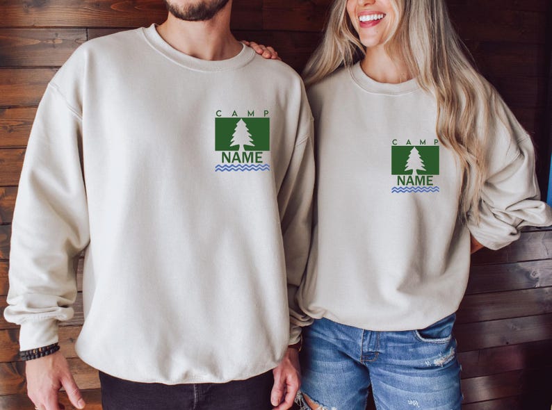 Puede incluir: Dos sudaderas beige. Cada una tiene un dise&ntilde;o gr&aacute;fico cuadrado verde con un &aacute;rbol blanco, la palabra "CAMP" arriba y "NAME" con l&iacute;neas onduladas azules debajo. Las sudaderas son de manga larga y las usan dos personas.